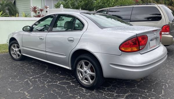 A silver four-door Oldsmobile Alero sedan seen from the rear left, parked on cracked asphalt with a tan minivan behind it.
