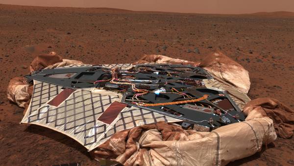 A dark metal Mars lander with tangled orange cabling, surrounded by crumpled cream-colored airbags with dark grid lines, on rust-red ground strewn with small rocks under a dusty orange sky.