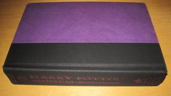 A hardcover book lying on a wooden surface, its plain purple cover facing up and its black cloth spine, printed in pink with the title Harry Potter and the Half-Blood Prince, toward the viewer.
