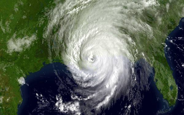 A satellite view of the northern Gulf of Mexico coast with the white spiral cloud mass of Hurricane Katrina and its small eye close to the shoreline, with green land to the north and dark Gulf waters to the south.