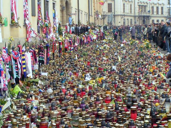 Thousands of red, amber and white memorial candles and bunches of yellow flowers covering a street in front of a pale building hung with football scarves, with a crowd standing at a distance.
