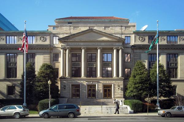 A neoclassical stone building with a columned entrance and a pediment lettered National Geographic Society, flagpoles on either side and cars parked at the curb.