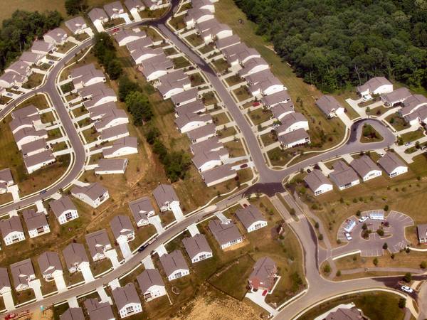 Aerial view of curving suburban streets lined with single-family houses with grey roofs, woodland at the upper right and a small parking circle at the lower right.