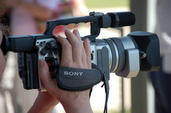 A silver and black Sony camcorder with a shotgun microphone above a long lens hood, held to the eye by a person whose hands and a sliver of cheek and hair show at the left edge.