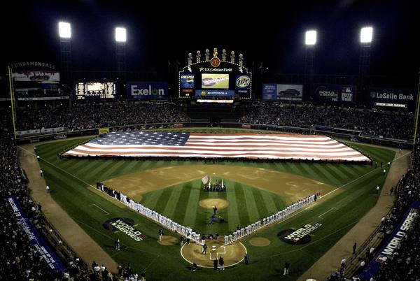 Night view from the upper deck of a packed baseball stadium, with a huge American flag spread across the outfield and the two teams lined up along the baselines beneath bright floodlights and a lit scoreboard.