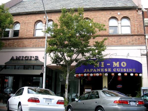 A two-story brick building with arched upper windows above a pizzeria and a Japanese restaurant, with a small tree and two parked cars in front.