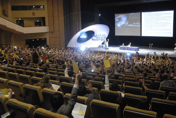 A large auditorium seen from behind the seats, with many delegates holding up yellow voting cards, a stage with a table and a swirling ringed IAU logo, and a projection screen showing the text of a resolution.