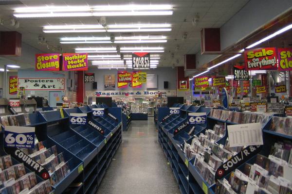 An aisle between blue shelving racks of CDs in a record store, with hanging Store Closing, Going Out of Business and 60% off signs and many empty shelves.