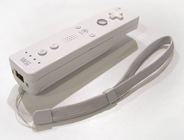 A white Wii Remote with its cross-shaped direction pad, round A button and numbered buttons lies at an angle on a white surface, with its grey fabric wrist strap looped in front of it.