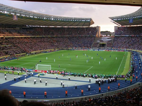 Wide view from the stands of a floodlit football stadium at dusk, with players in blue and white small on the green pitch, a blue running track around it and crowded stands under the roof.