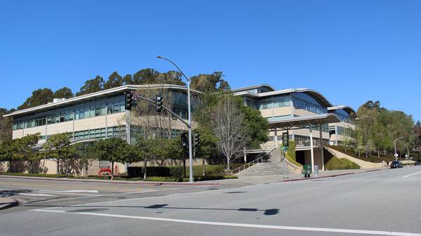 A low-rise office building with curved rooflines, glass windows and trees, seen from a street corner under a clear blue sky.