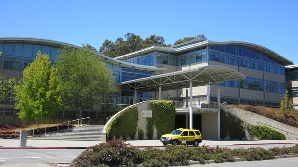 A curved glass-and-stucco office building with a canopy over a stairway entrance, a yellow taxi in front, shrubs in the foreground and a clear blue sky.