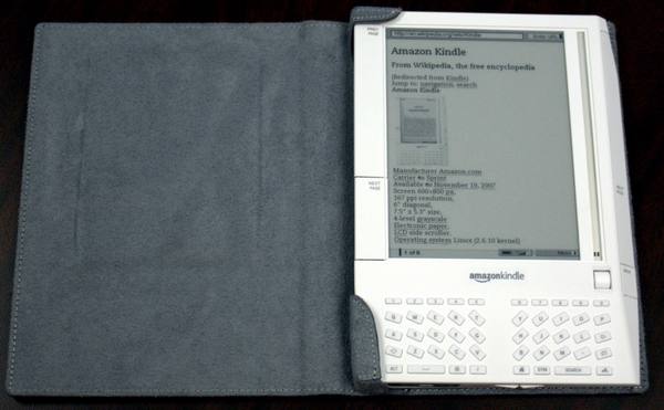 A white first-generation Kindle e-reader lying open on a gray suede cover, its screen showing a page of text and its keyboard visible below the screen.