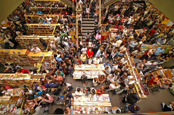 A view from an upper floor down into a bookstore packed with people standing between wooden shelves and display tables.