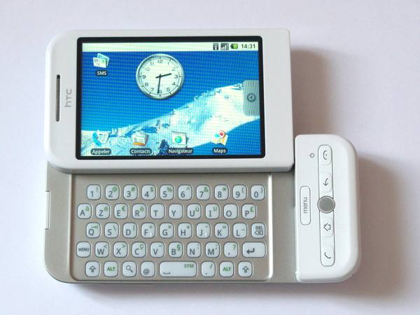 A white HTC smartphone lying flat with its screen slid sideways to reveal a full keyboard, the screen showing a blue Android home screen with a clock and French-language icons.