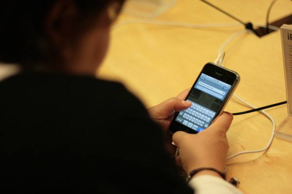 Two hands hold an original iPhone showing a web browser with the on-screen keyboard, while the dark silhouette of the user's head and shoulder fills the left of the frame.