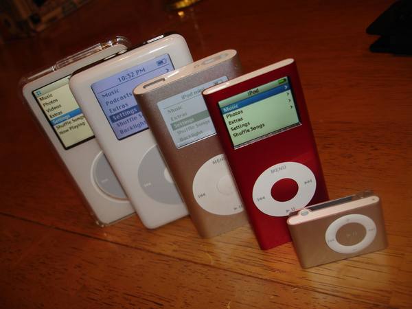 Five Apple iPod music players of different sizes standing in a row on a wooden table, with the small iPod shuffle in front and the red iPod nano beside it.