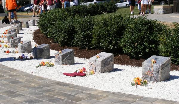 A curved row of rough gray stone blocks on white gravel, with orange and yellow flowers and a folded maroon cloth left beside them, in front of dark green shrubs; a few visitors are partly visible at the top edge.