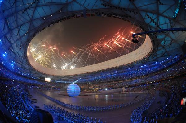 Inside the Beijing National Stadium at night: fireworks burst above the open roof ring, a glowing globe stands on the field, and performers and spectators fill the stands.