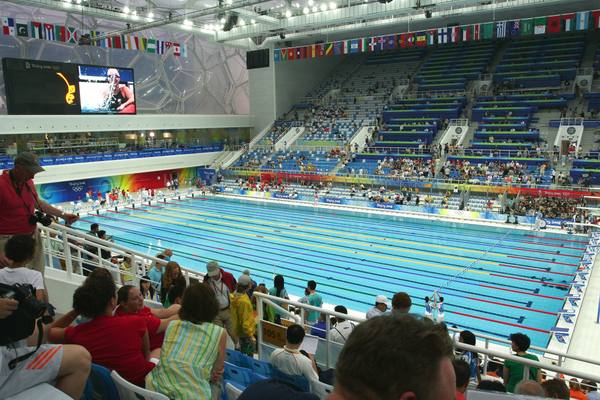 The interior of a large indoor swimming arena with a long pool in the center, stands of blue seats, national flags hanging along the walls, a big video screen at upper left and spectators in the foreground.