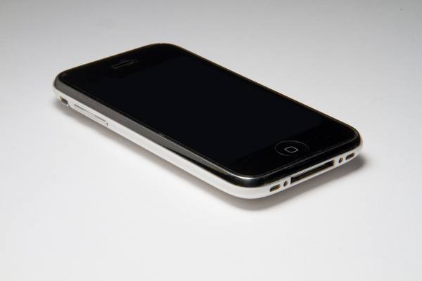 An iPhone 3G lies at an angle on a plain white background with its black screen switched off, its round home button at the lower right and its white plastic edge and dock connector in front.