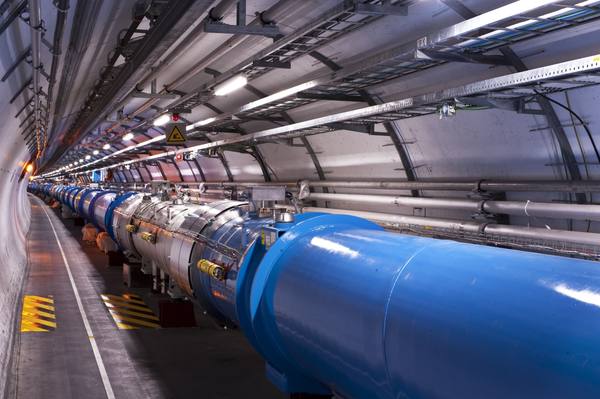 A long underground tunnel curving into the distance, with a large blue cylindrical machine along the right wall and cable trays, pipes and lights overhead.