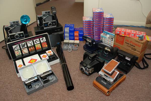 Several Polaroid instant cameras, including folding pack-film models and an SX-70 in a tan leather case, with stacks of Polaroid 600, 664 and 669 film boxes and a box of flash cubes arranged on a carpet.