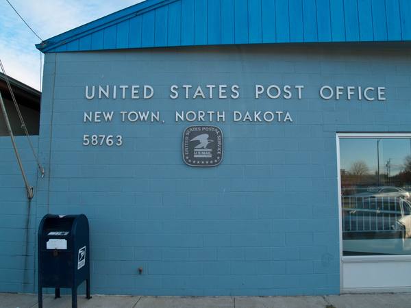 A light blue cinder-block post office wall with raised lettering reading United States Post Office, New Town, North Dakota 58763, a Postal Service eagle plaque, and a dark blue collection box on the sidewalk.