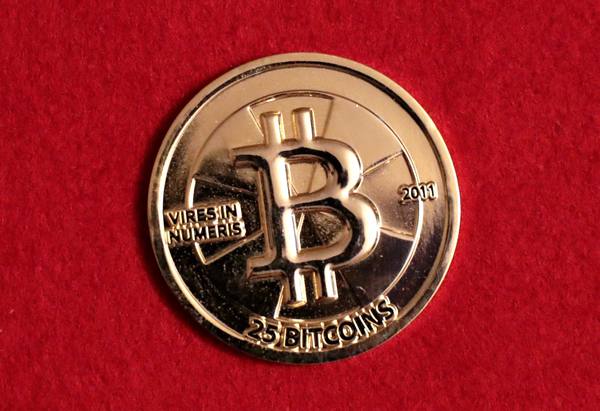 A round gold-colored coin stamped with a large letter B, the date 2011, the words 25 Bitcoins and the Latin motto Vires in Numeris, lying on red cloth.