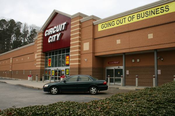 A brick Circuit City store with a red entrance tower, a large yellow Going Out of Business banner and a dark sedan in the parking lot.