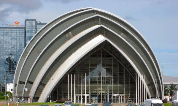 A building made of overlapping curved silver-grey ribs arches over a tall glass entrance front under a partly cloudy sky, with a glass-fronted tower behind it on the left and parked vehicles along the bottom edge.