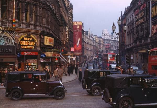 A color street scene of Shaftesbury Avenue with black taxis in the foreground, red double-decker buses, pedestrians and illuminated shop and cinema signs.