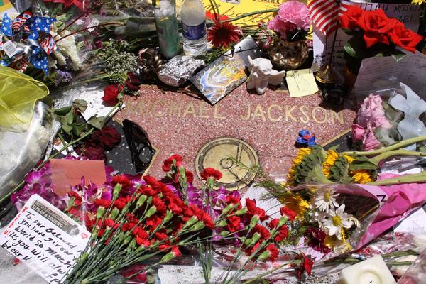 A pink terrazzo Hollywood Walk of Fame star engraved with the name Michael Jackson, surrounded by red carnations, sunflowers, roses, handwritten notes, a small white cherub figure and a small American flag.