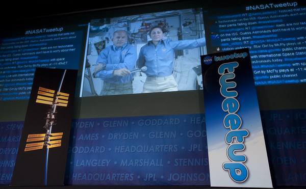 A dark stage with a large video screen showing two astronauts in blue shirts aboard the space station, flanked by screens of scrolling #NASATweetup posts, a space station banner at left and a tall NASA Tweetup banner at right.