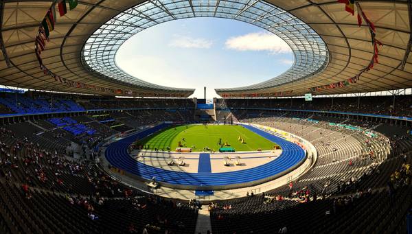 A wide panoramic view into the Berlin Olympiastadion: a blue running track around a green infield, under the oval opening of the roof with national flags hanging from its edge, and spectators scattered across the stands.
