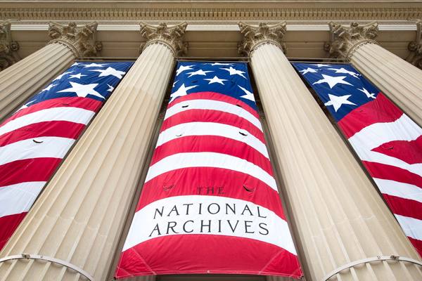 Looking up at tall fluted stone columns hung with large red, white and blue flag-pattern banners, the center one reading The National Archives.