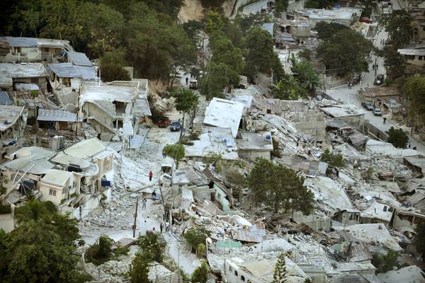 Aerial view of a hillside neighbourhood where many concrete houses have collapsed into grey rubble along a street, with trees around them and a few tiny figures walking in the distance.
