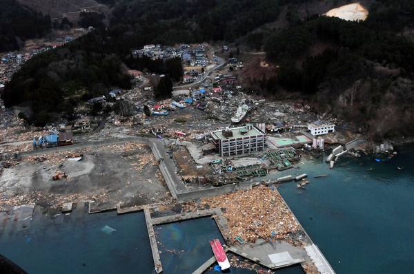 Aerial view of a coastal town reduced to fields of debris and gutted buildings beside a harbour, with wooded hills behind it.