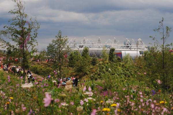 Wildflower meadows and young trees in the foreground, with crowds of visitors on the paths and the white-roofed Olympic Stadium in the distance under a cloudy sky.