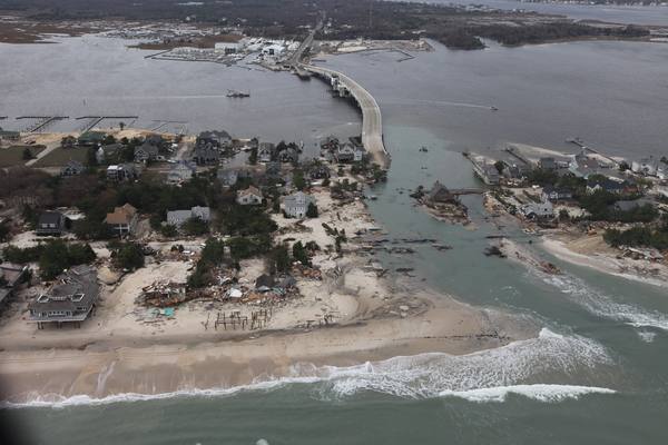 Aerial view of a barrier-island town where the sea has cut a channel through the beach next to a bridge, leaving damaged and missing houses scattered across the sand.