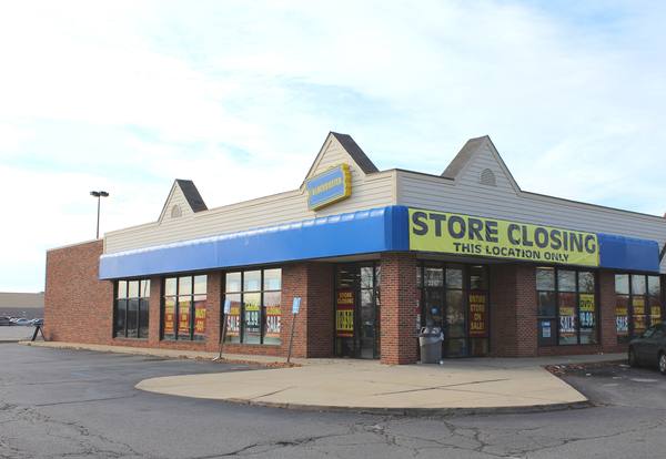 A brick video-rental store with a blue awning and a large yellow 'Store Closing – This location only' banner, sale signs in the windows and a nearly empty parking lot.