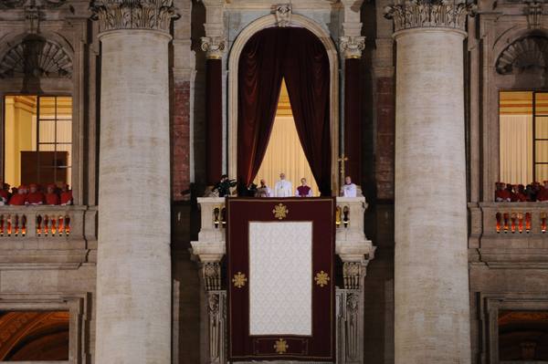 A small white-robed figure stands on a central balcony hung with a crimson banner, framed by tall stone columns, with cardinals in red on the side balconies.