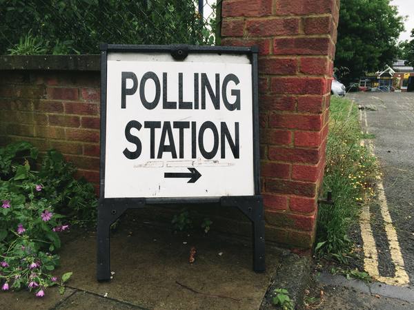 A white sandwich-board sign reading 'Polling Station' with a black arrow, propped against a brick gatepost beside a pavement with double yellow lines and weeds.