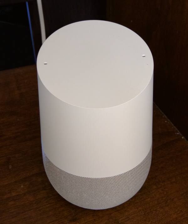 A white cylindrical voice-assistant speaker with a slanted top and a grey fabric base, sitting on a wooden surface.