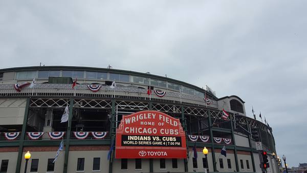 The red marquee of a ballpark reading 'Wrigley Field, Home of Chicago Cubs' and 'Indians vs. Cubs, World Series Game 4', with red, white and blue bunting along the stands under a grey sky.