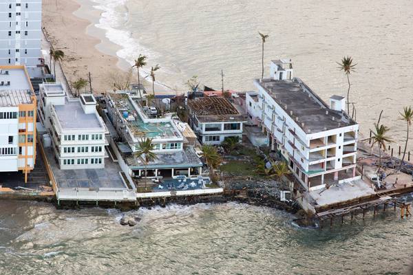 Aerial view of beachfront apartment buildings with torn roofs and broken balconies, palm trees stripped of leaves, and waves washing against a damaged seawall.