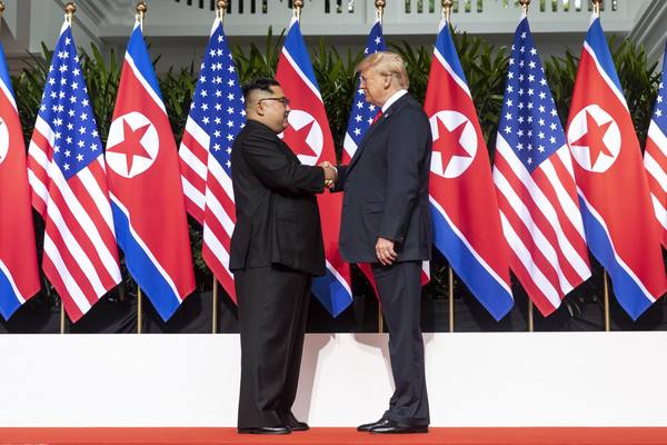 Two men in dark suits shake hands on a red carpet in front of a row of alternating U.S. and North Korean flags.